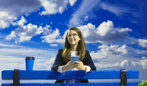 Smiling woman holding mobile phone, looking at clouds overhead.
