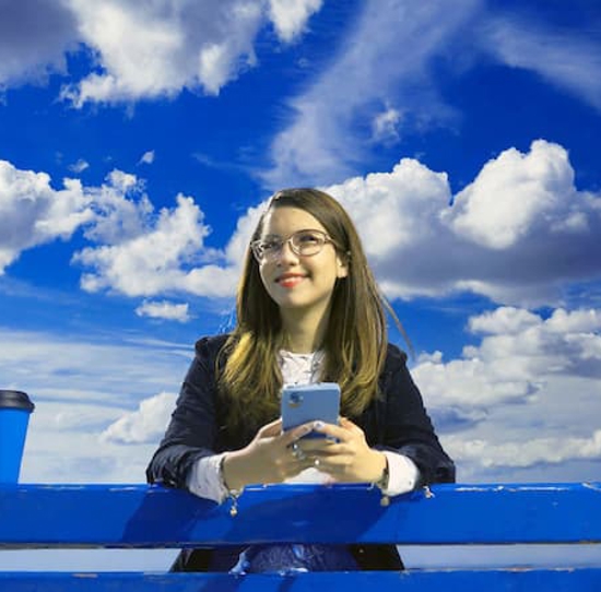 Smiling woman holding mobile phone, looking at clouds overhead.
