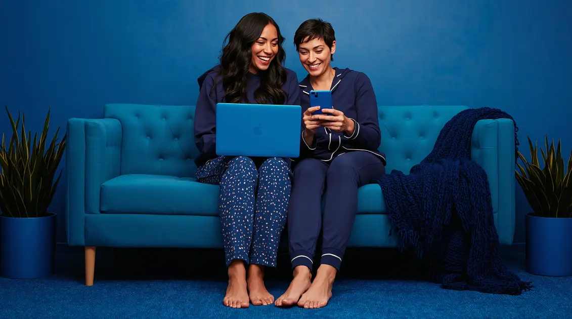 2 smiling women sitting on couch together and looking at mobile devices.